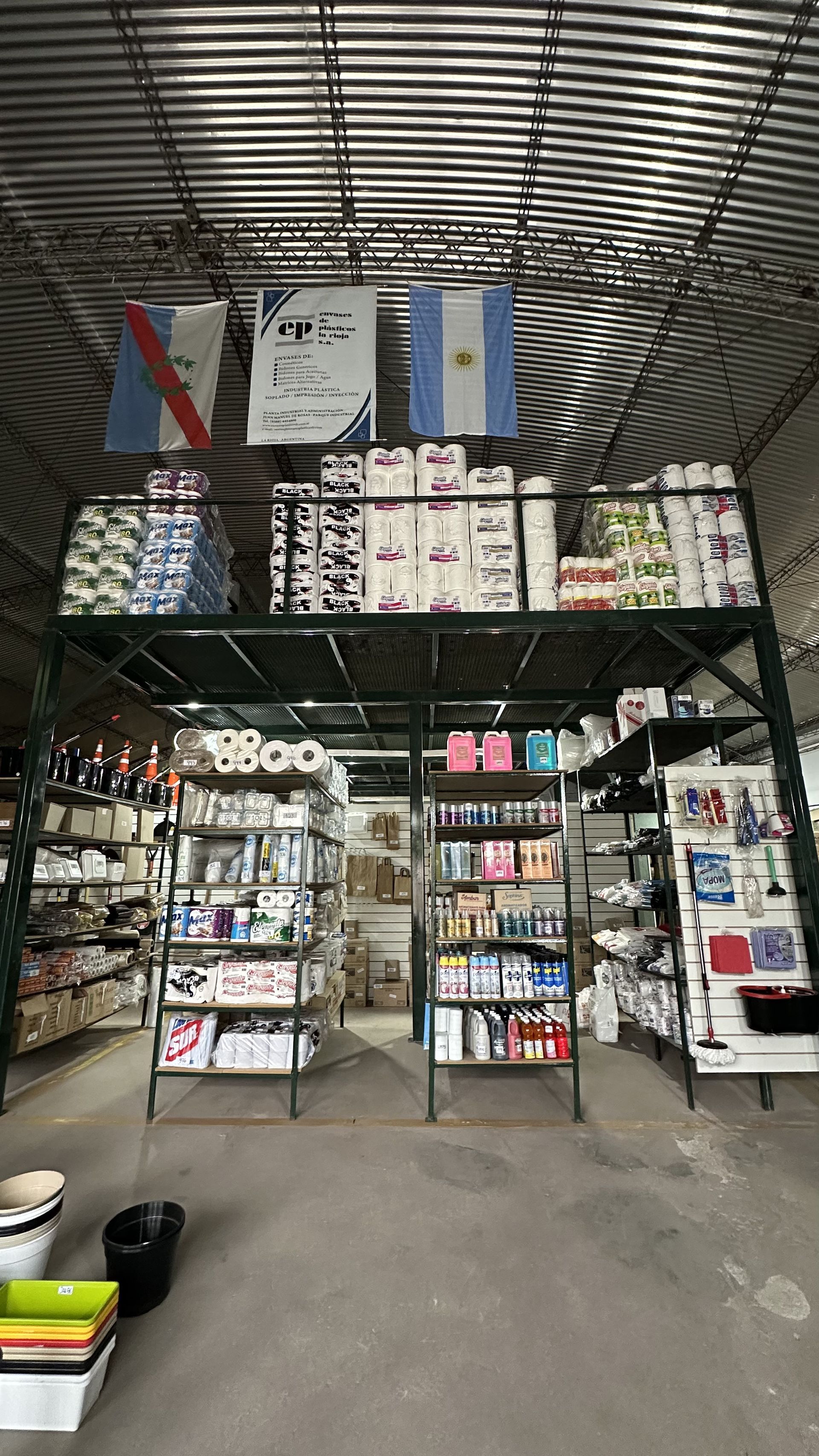 Pequeña tienda abierta por delante, con estanterías llenas de productos bajo un techo de metal, vista desde la entrada.