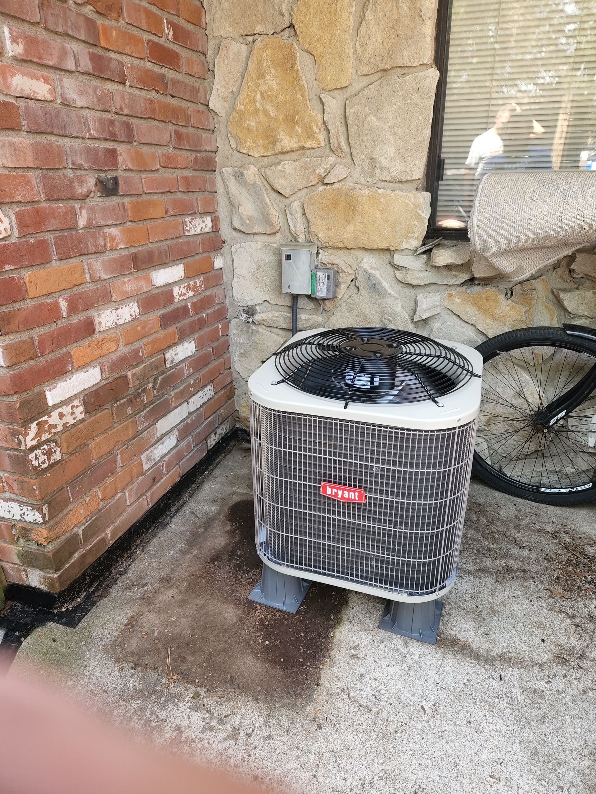 An air conditioner is sitting in front of a brick wall next to a bicycle.