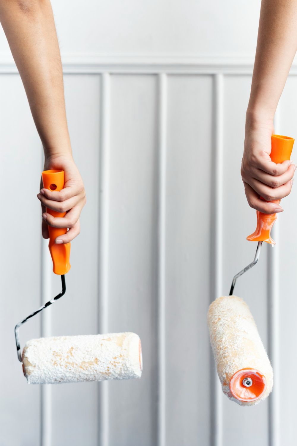 Hands holding paint rollers with white paint against a light gray wall.