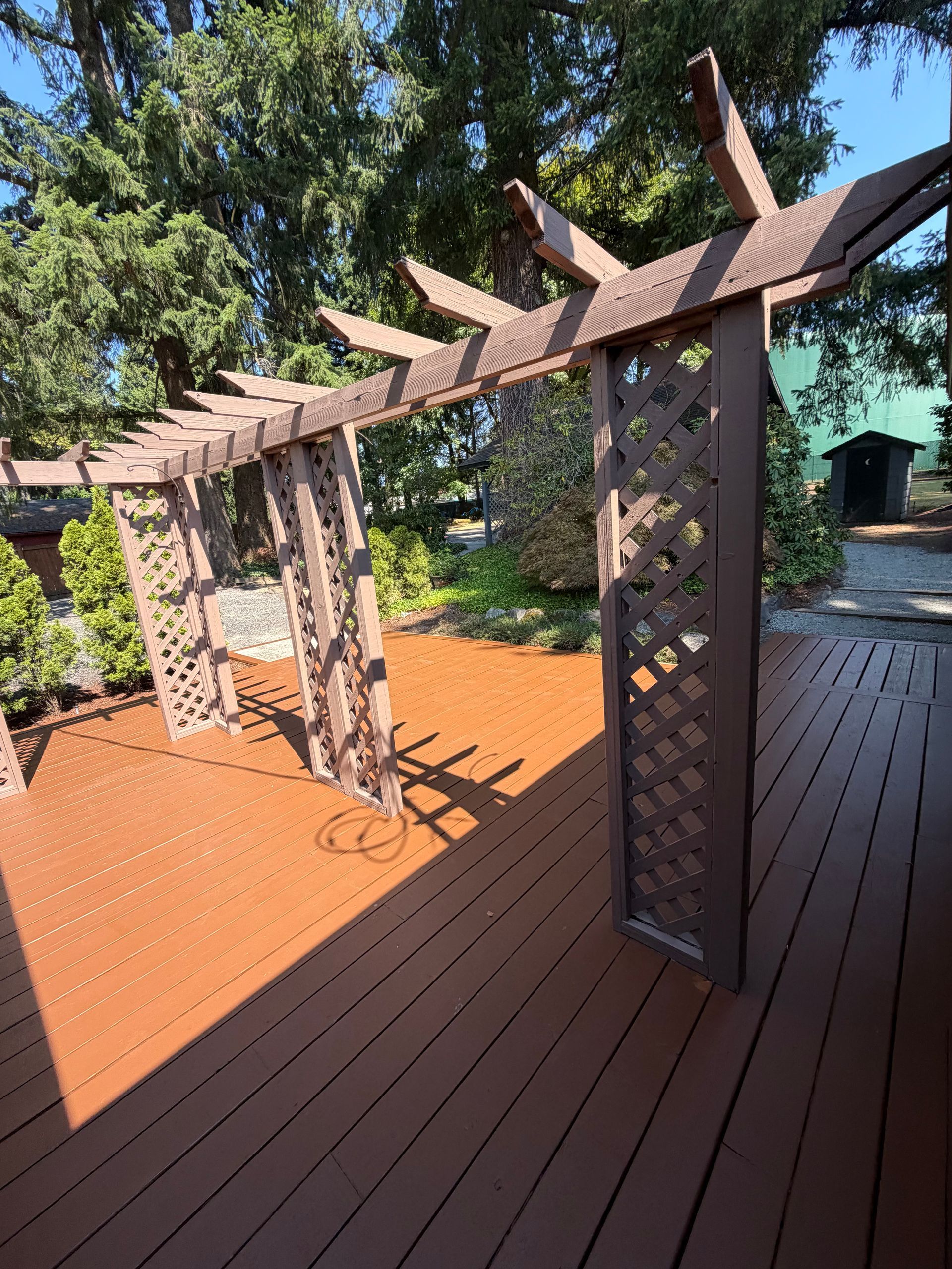 Wooden pergola with lattice posts on a sunlit deck, surrounded by trees and greenery