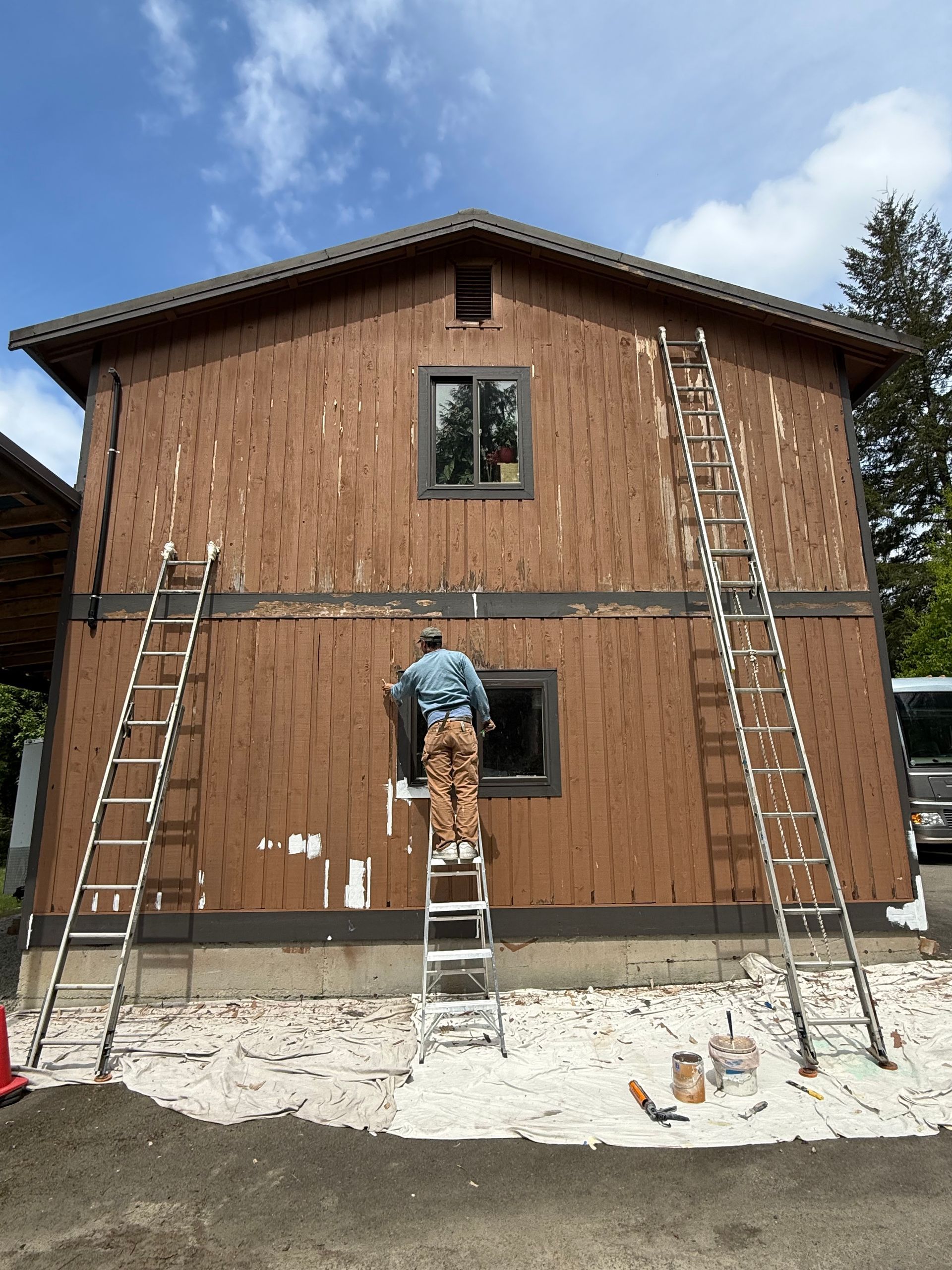 Worker on a ladder painting a two-story house with brown siding and a gray foundation