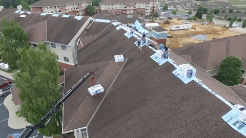 Roofers replacing shingles on a large brown-roofed building. Crane lifts materials.