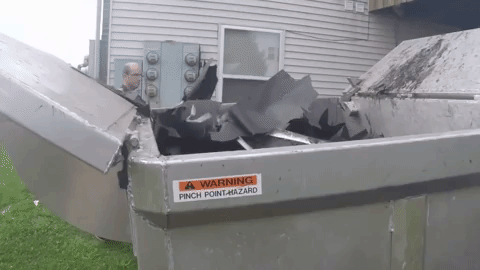 Man looks into a metal dumpster filled with black debris near a building with electrical boxes.