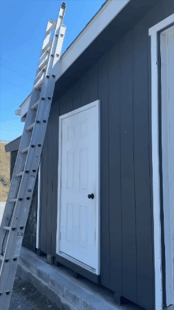 Ladder leaning against a gray building beside a white door under a bright blue sky