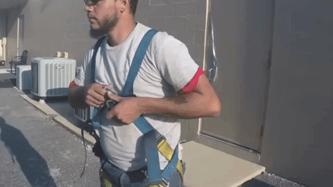 Man in a white shirt and safety harness buckles the chest strap. Outside.
