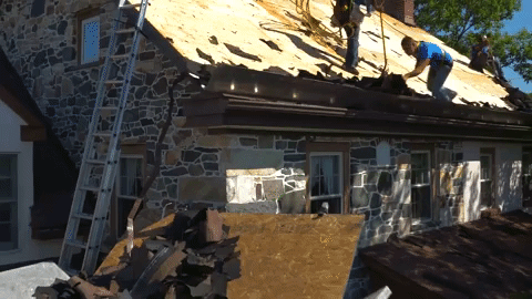 Roofers removing old shingles from a stone house roof. A ladder is leaning against the house.