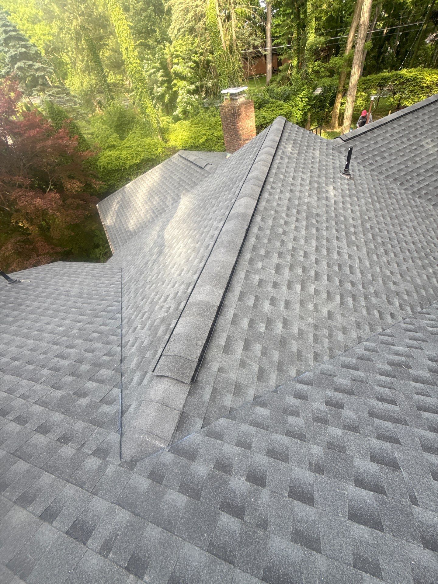 Gray shingled roof with a chimney, surrounded by green trees in sunlight