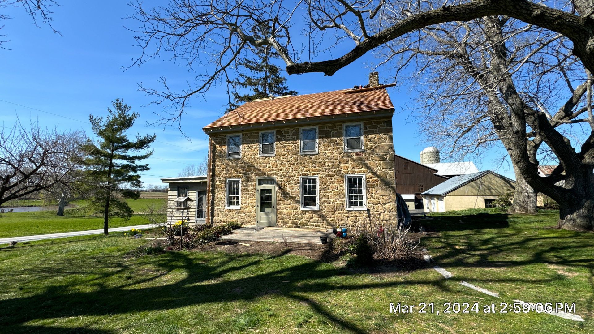 Stone house with red tile roof, on a grassy lawn. Large tree frames the building on sunny day.
