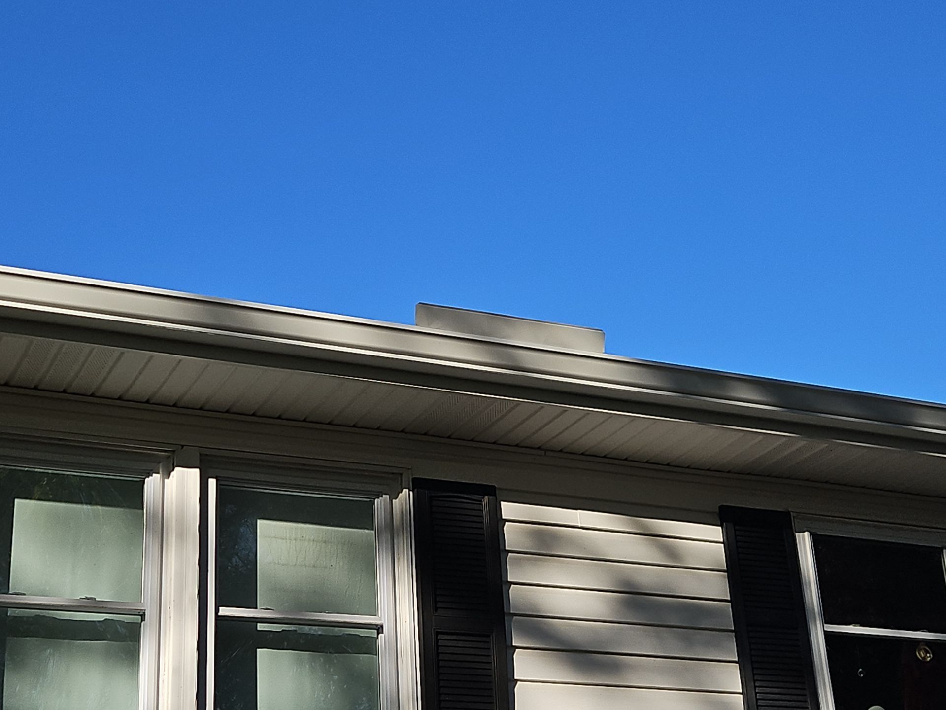 House siding and windows beneath a blue sky, with roof eaves and black shutters visible