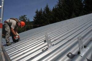 Roofer on a sloped metal roof, wearing a hard hat, cutting material near safety attachments, trees in background.