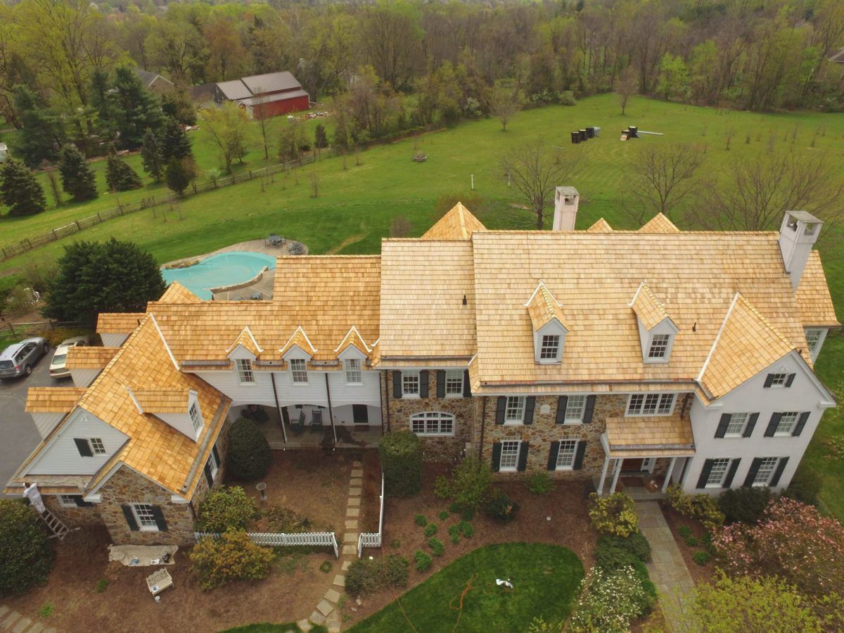 Aerial view of a large stone-and-white house with tan roof, surrounded by trees and lawn.