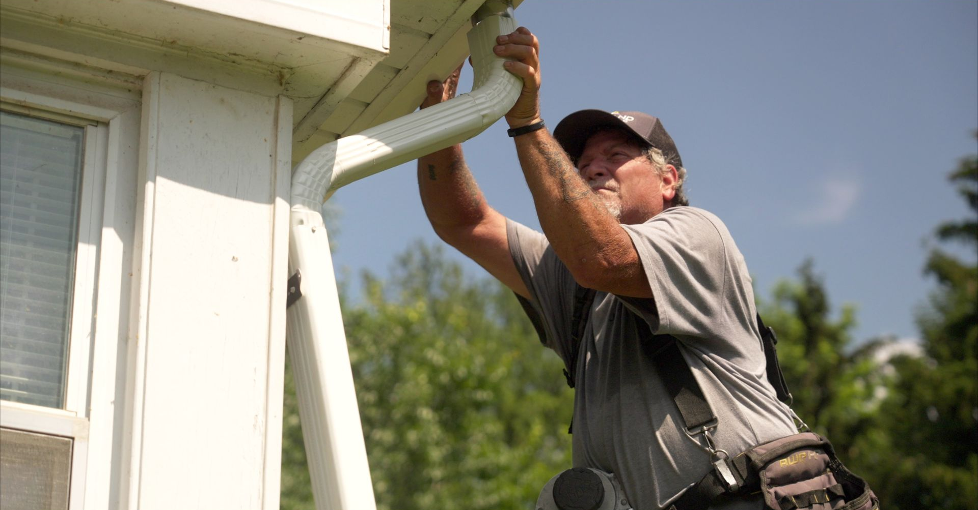 Man on a ladder installing a white gutter on a house, sunny outdoor setting.