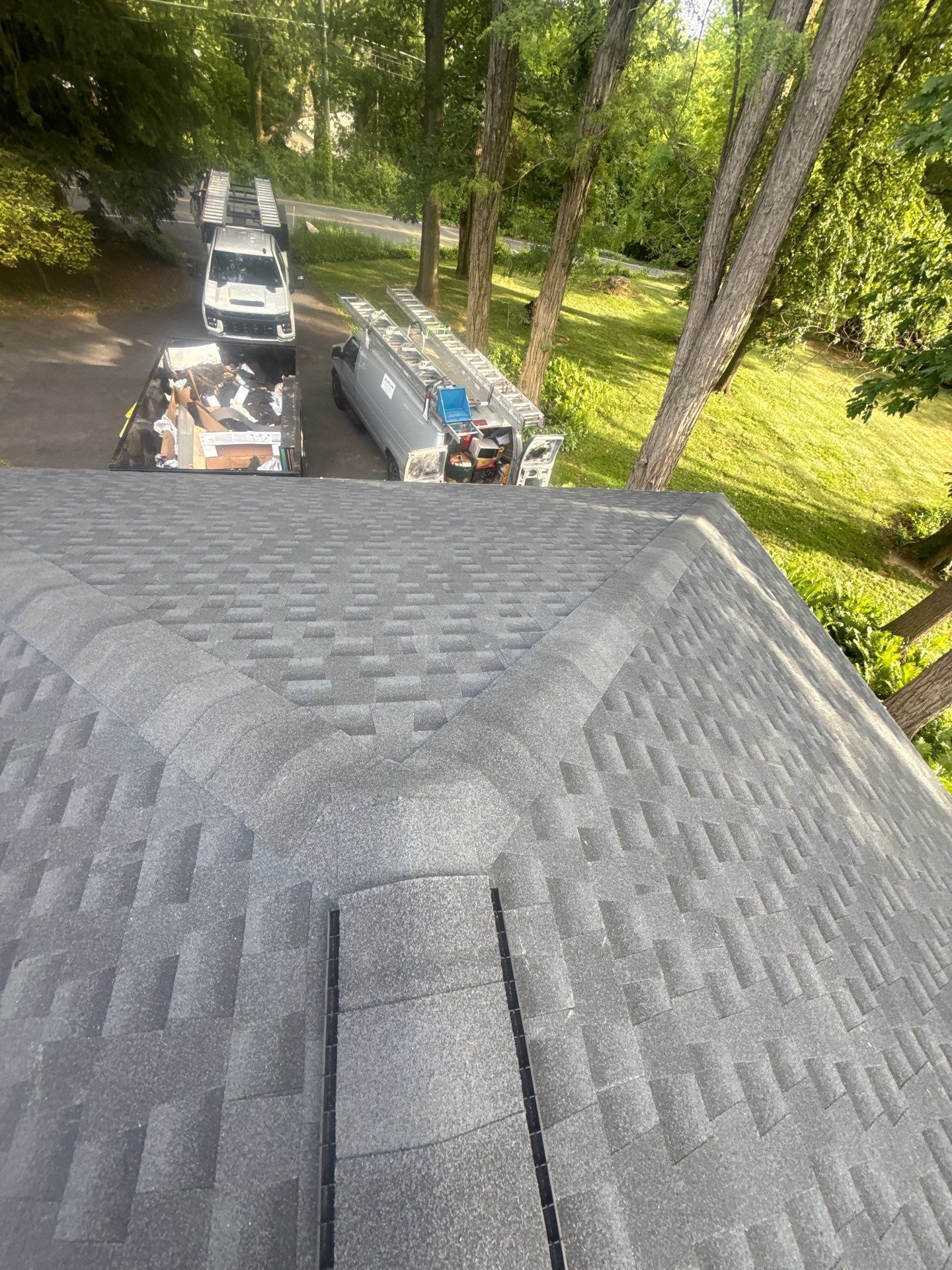 View from a shingled roof onto a wooded driveway with two parked vehicles and a small trailer below