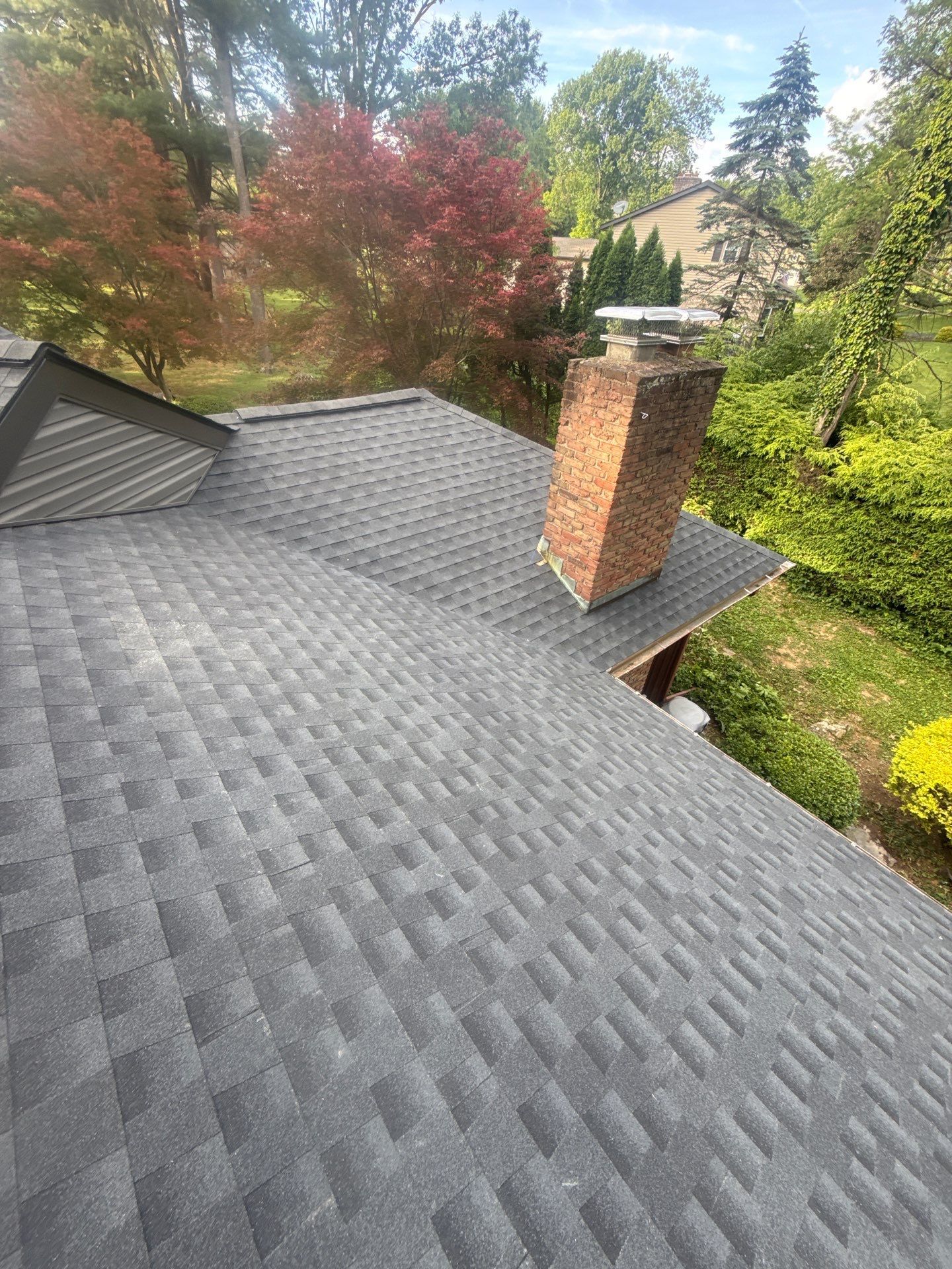 Rooftop with gray shingles and a brick chimney, overlooking green trees and a neighboring house