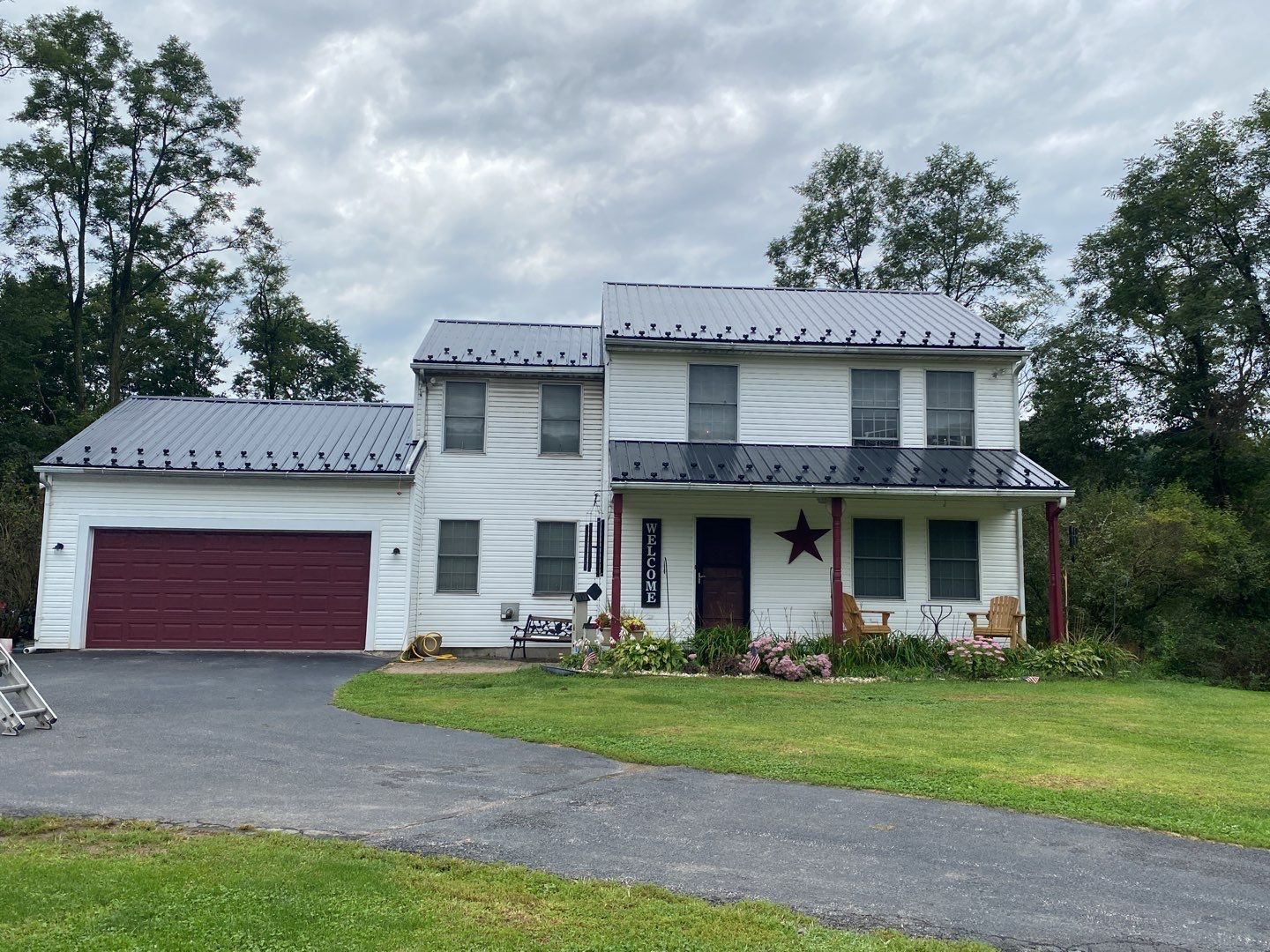 A white two-story house with a grey metal roof, a maroon garage door, and a front porch with chairs on a grassy lot.