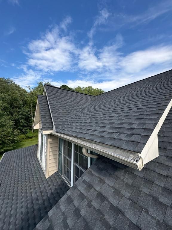 Gray shingled roof with dormer and white trim under a blue sky, viewed from above.