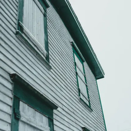 Side view of an old, weathered house with boarded-up windows. Gray siding, teal trim, overcast sky.