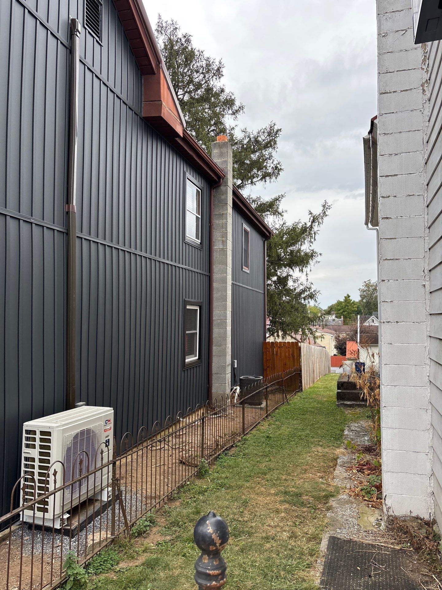 Narrow alley between black siding and white brick buildings, with a fence and air conditioner unit.