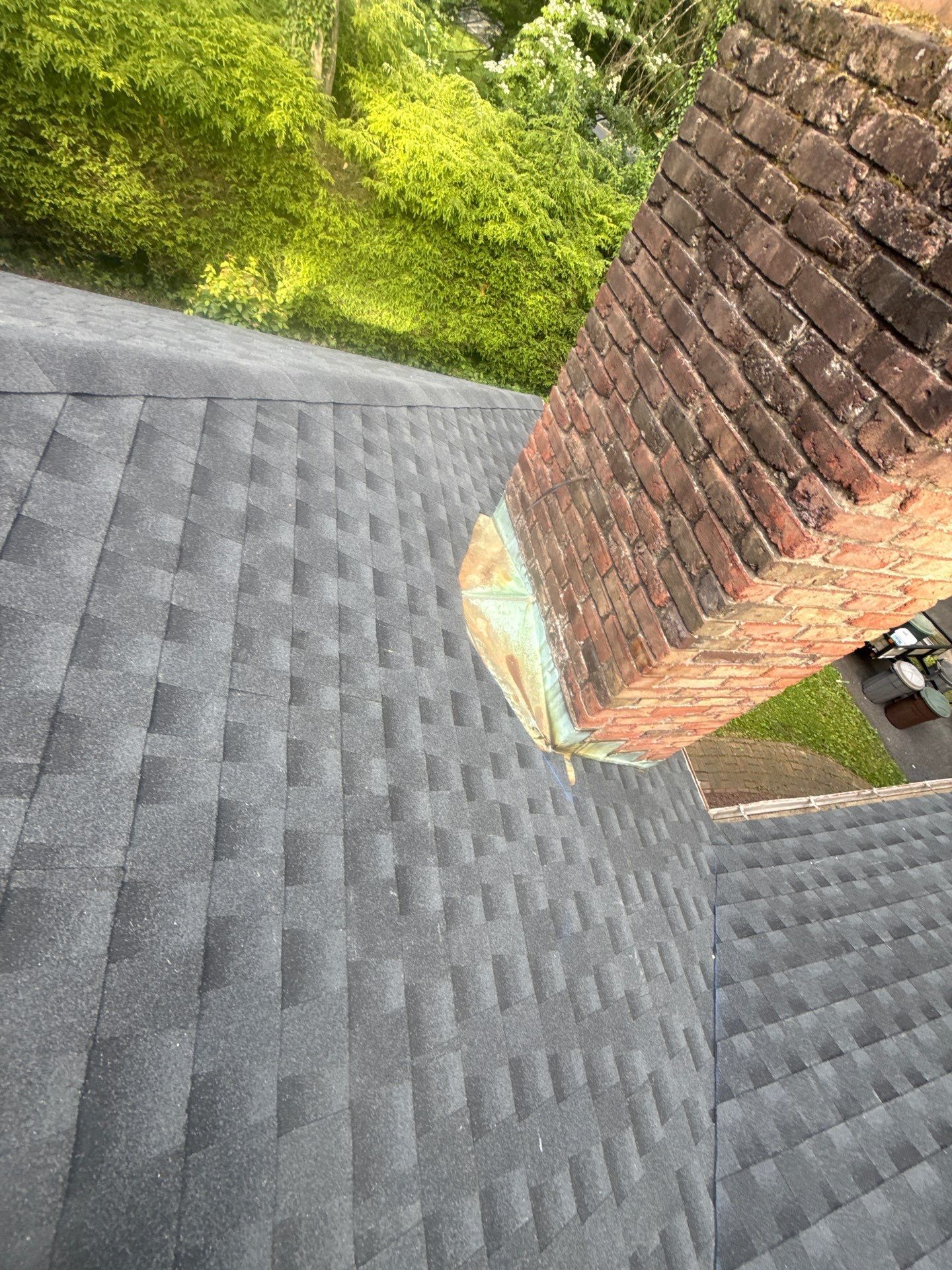 Gray shingled roof beside a brick chimney, with green trees below.