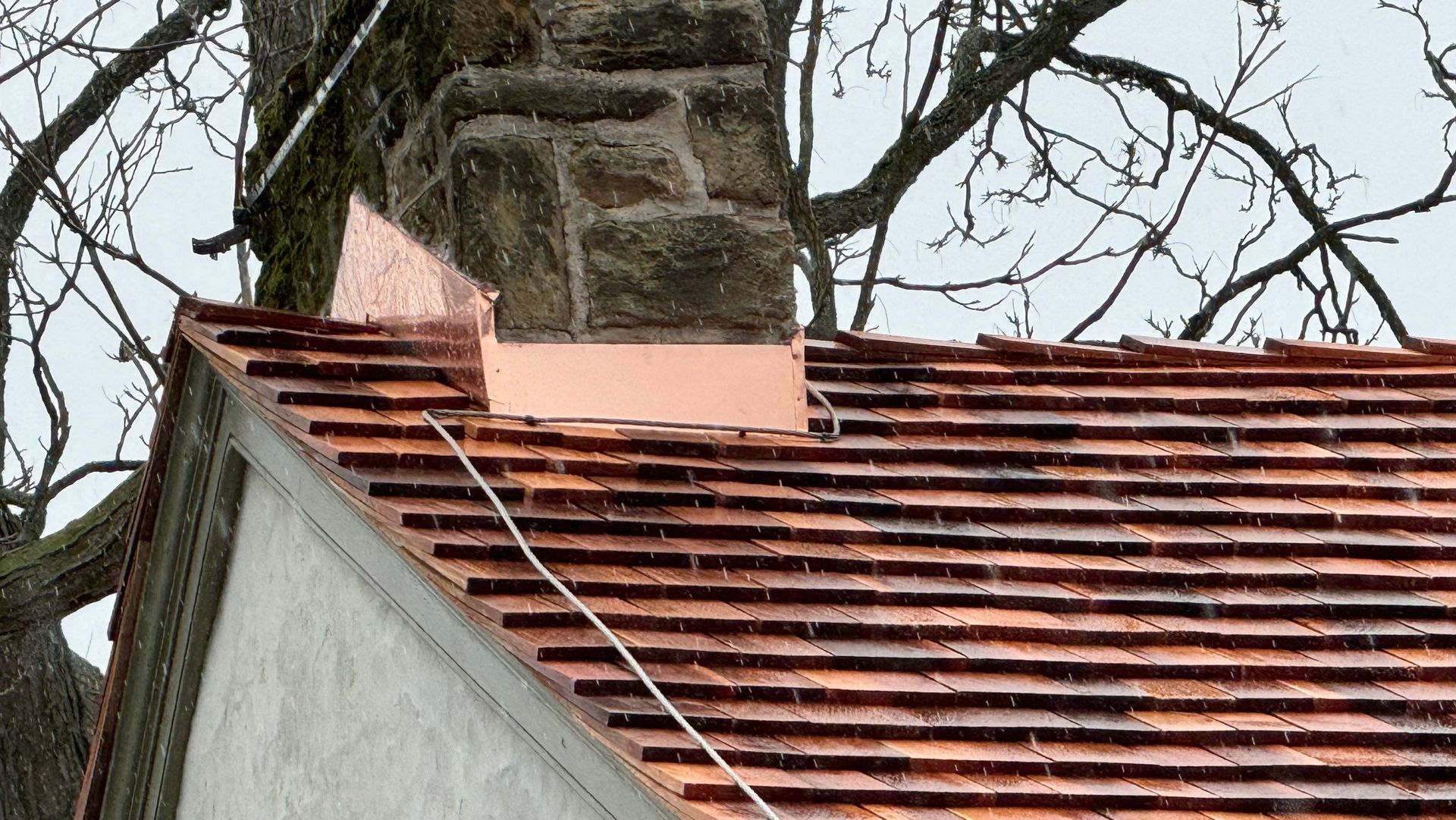 Copper flashing around a stone chimney on a red-tiled roof, with bare tree branches in the background.