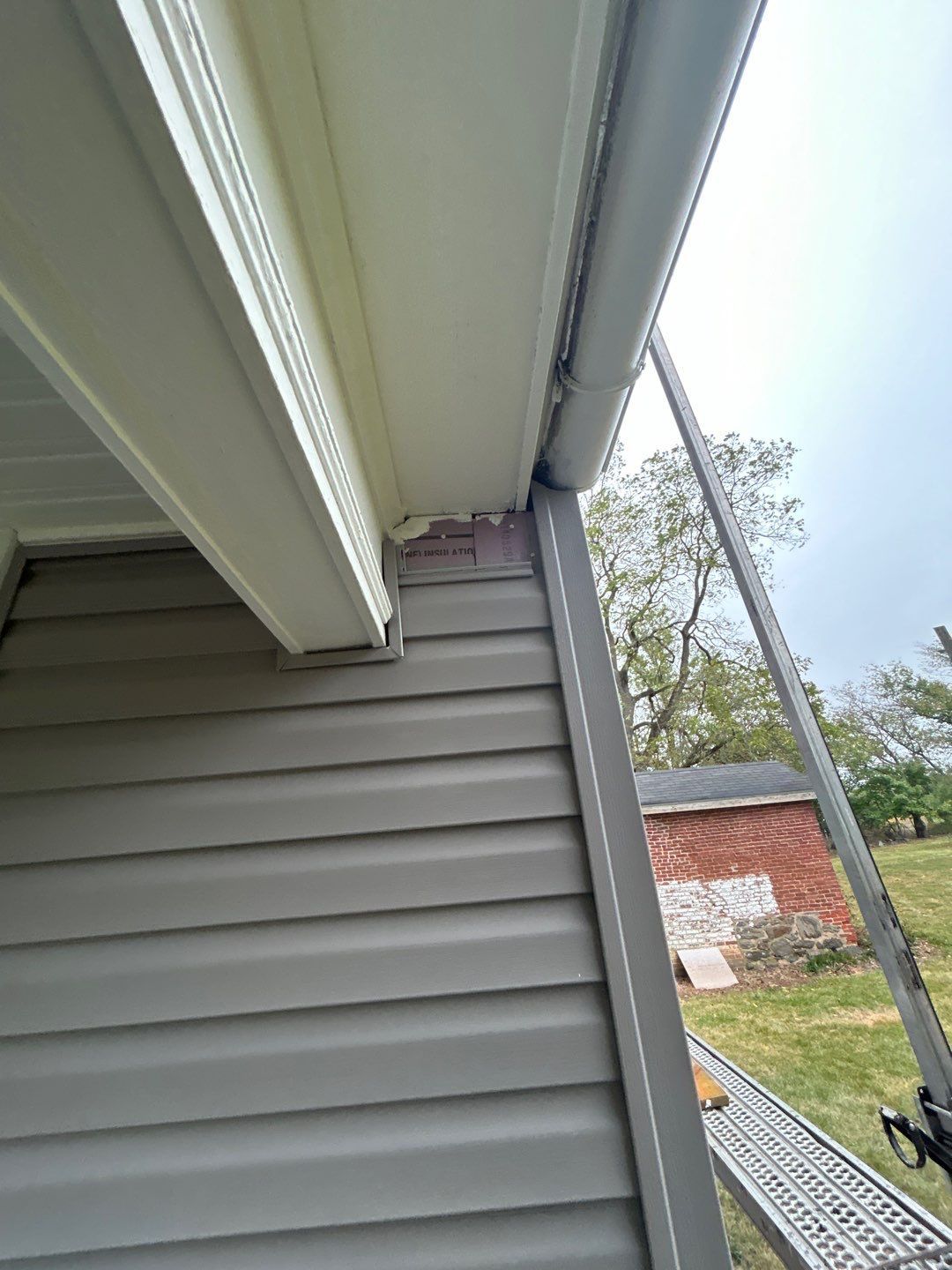 Gray house siding with white trim, gutter, and ladder in outdoor setting.