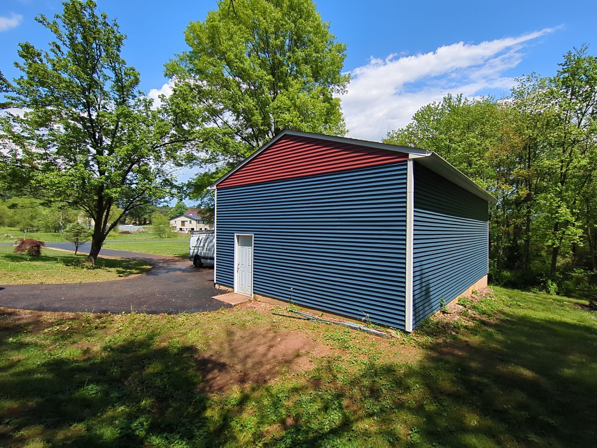 Blue and red barn with a white door, set in a yard with trees and a driveway on a sunny day.