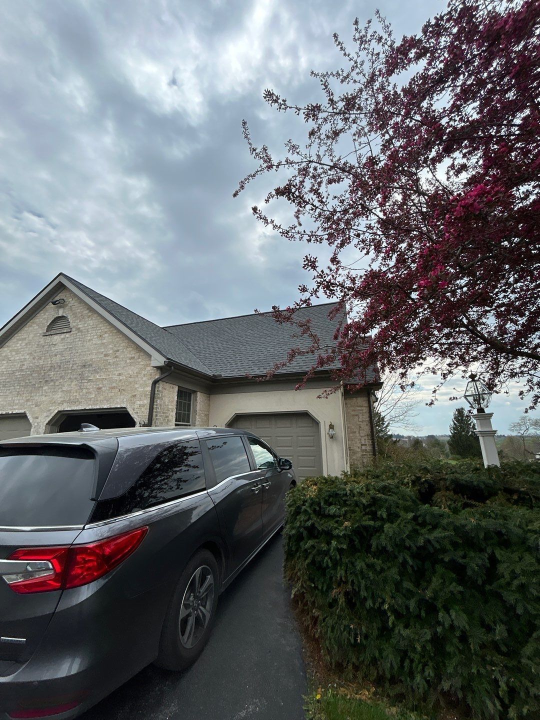 Dark SUV parked on a driveway in front of a tan brick house with a dark roof and a red-leaved tree.