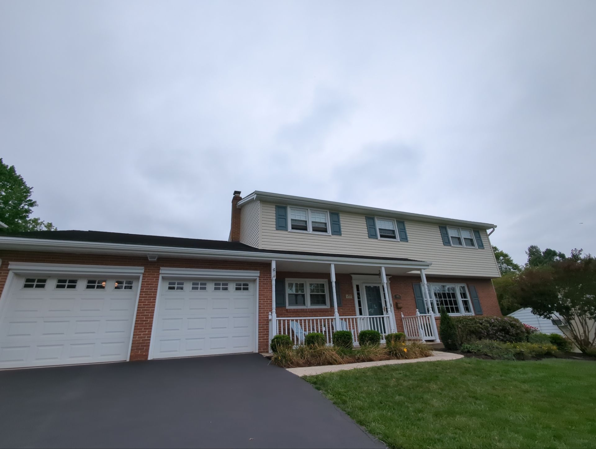 Two-story house with beige siding, brick facade, white garage doors, porch, and a cloudy sky.