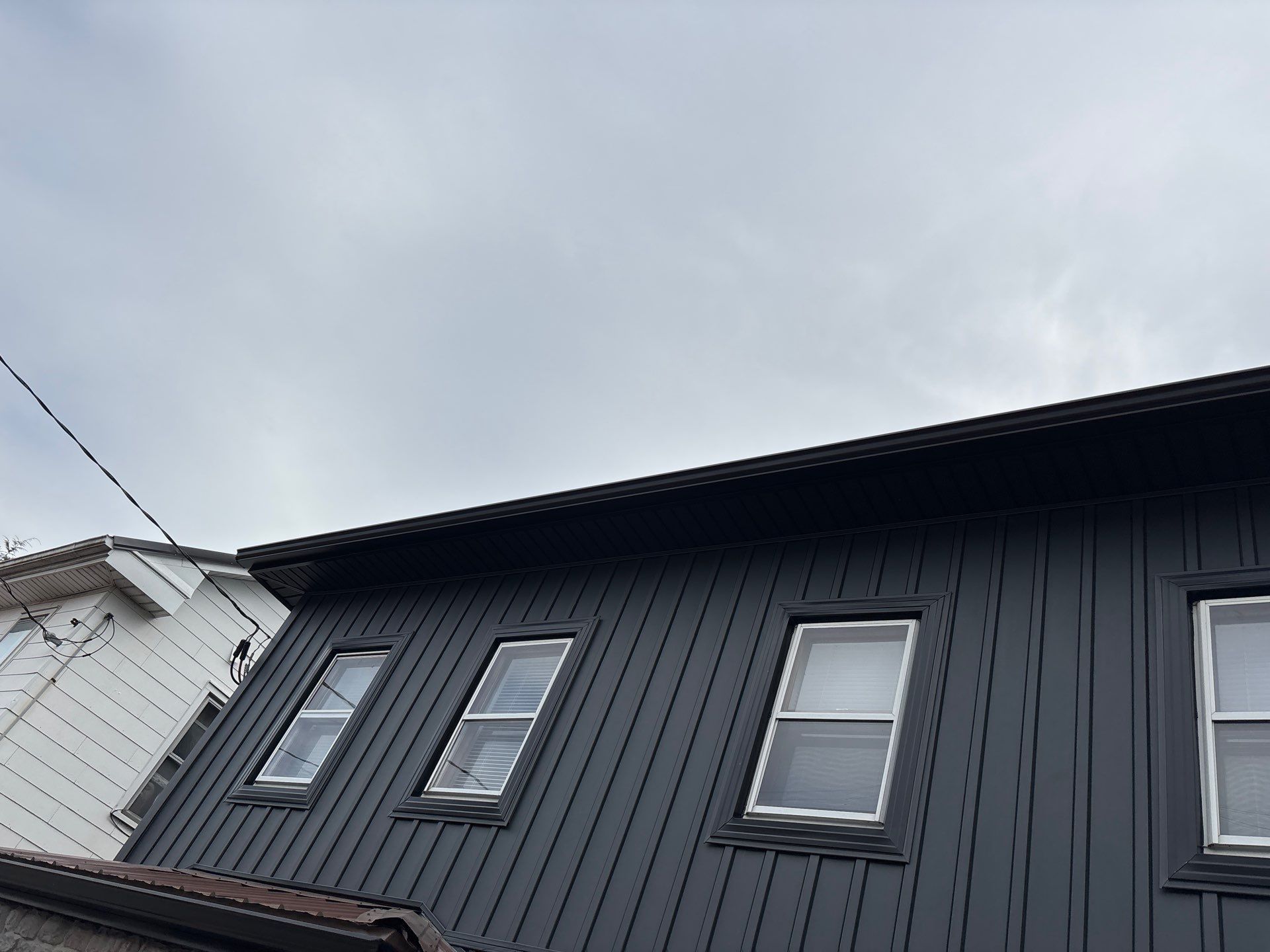 Dark gray house exterior with slanted roof and several windows under an overcast sky