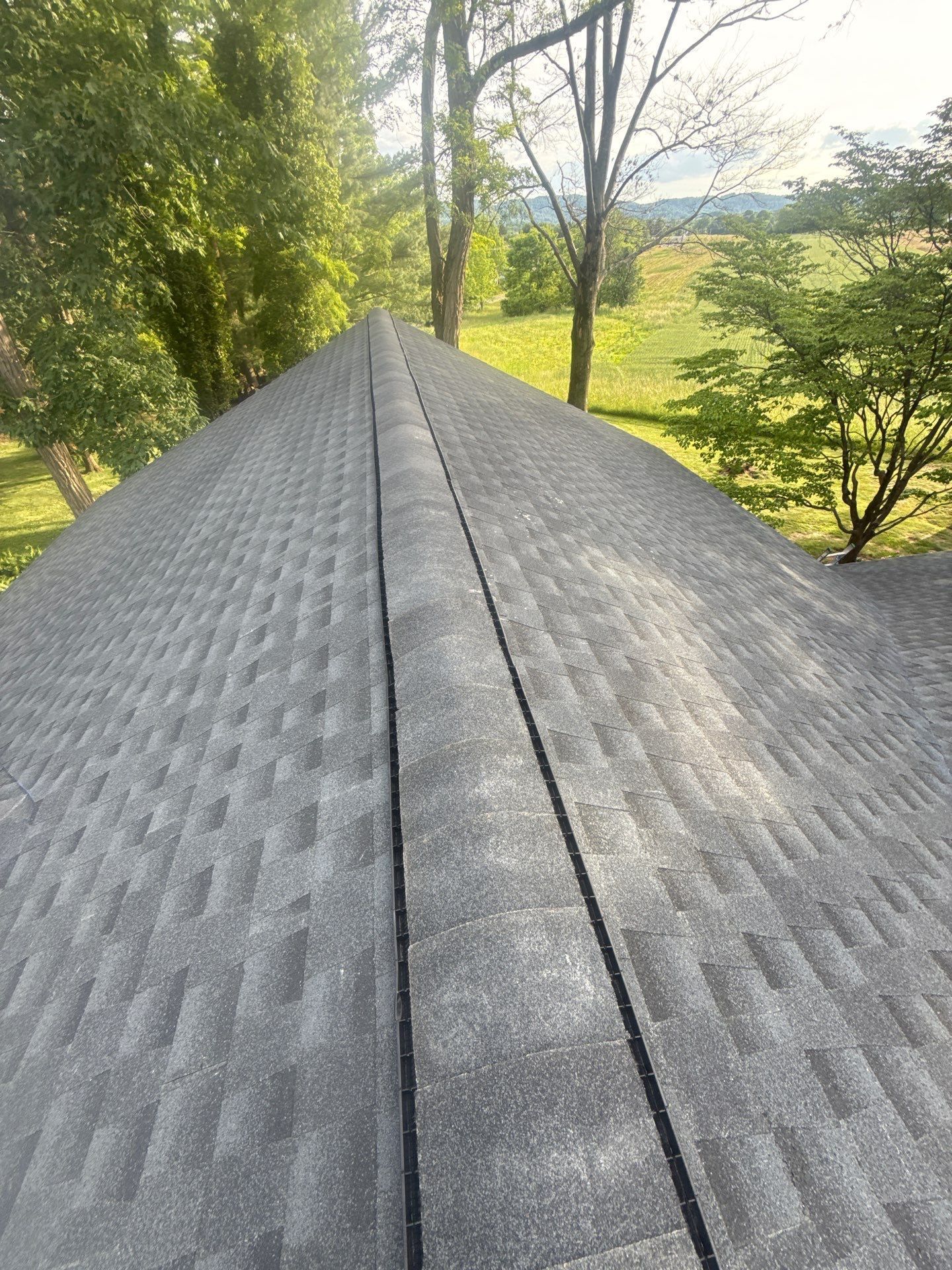 Roof ridge with gray shingles overlooking green trees and fields under a sunny sky