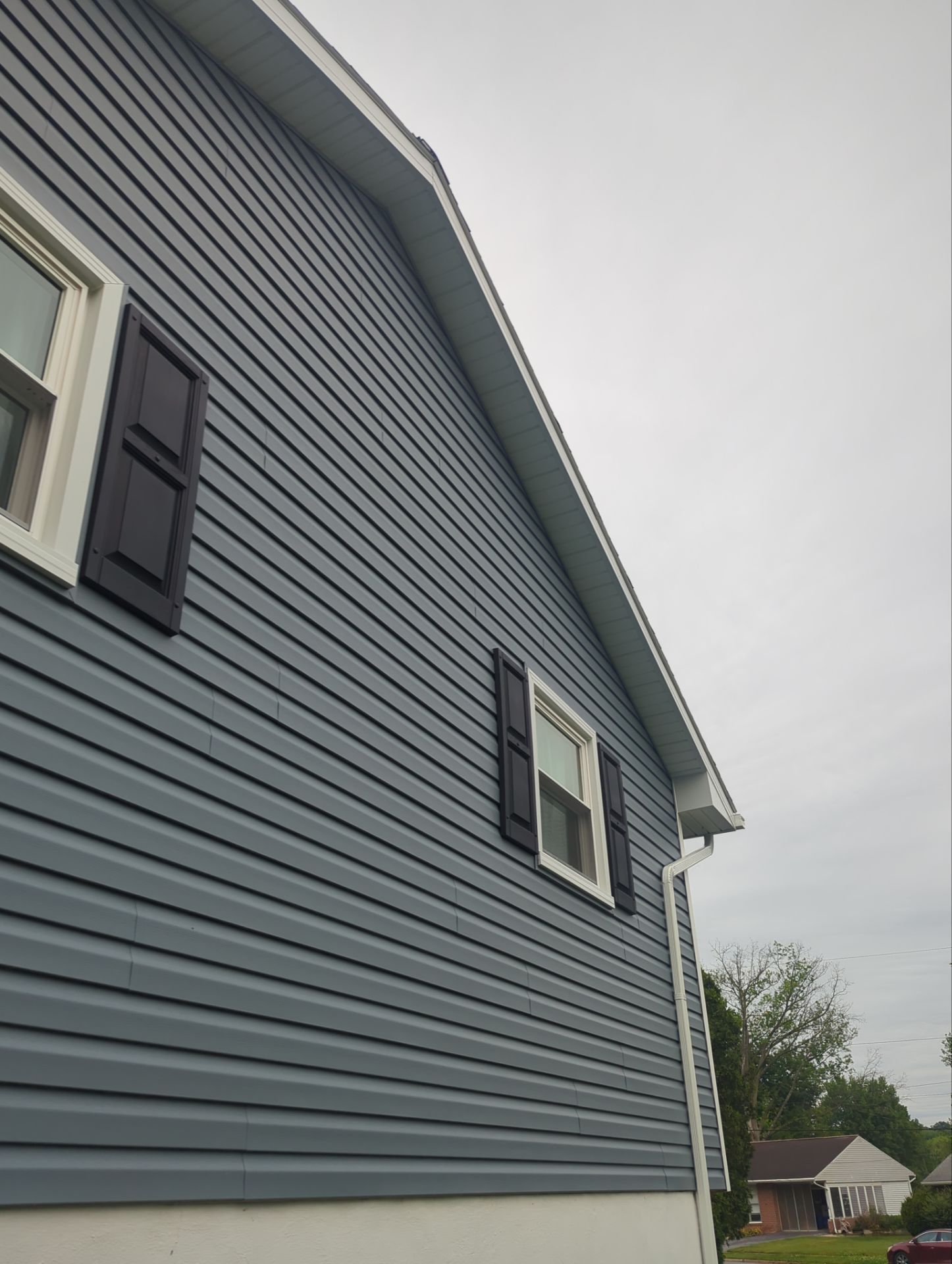 Two-story house with tan siding, black shutters, white porch with hanging plant, and a cloudy sky.