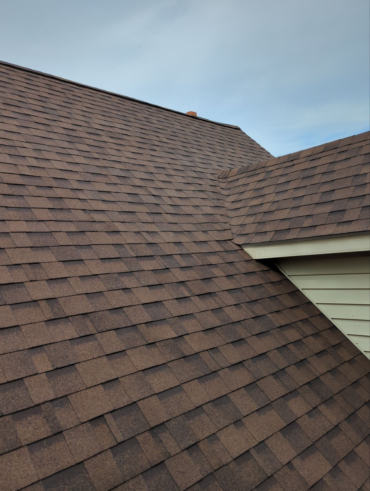 Brown shingle roof on a house under a pale blue sky, with a sloped section and white siding visible