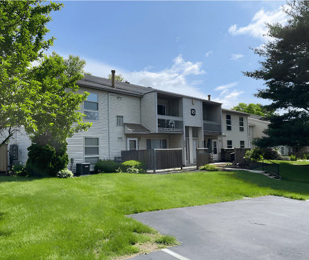 Two-story beige apartment building with balconies, green lawn, trees, and blue sky.