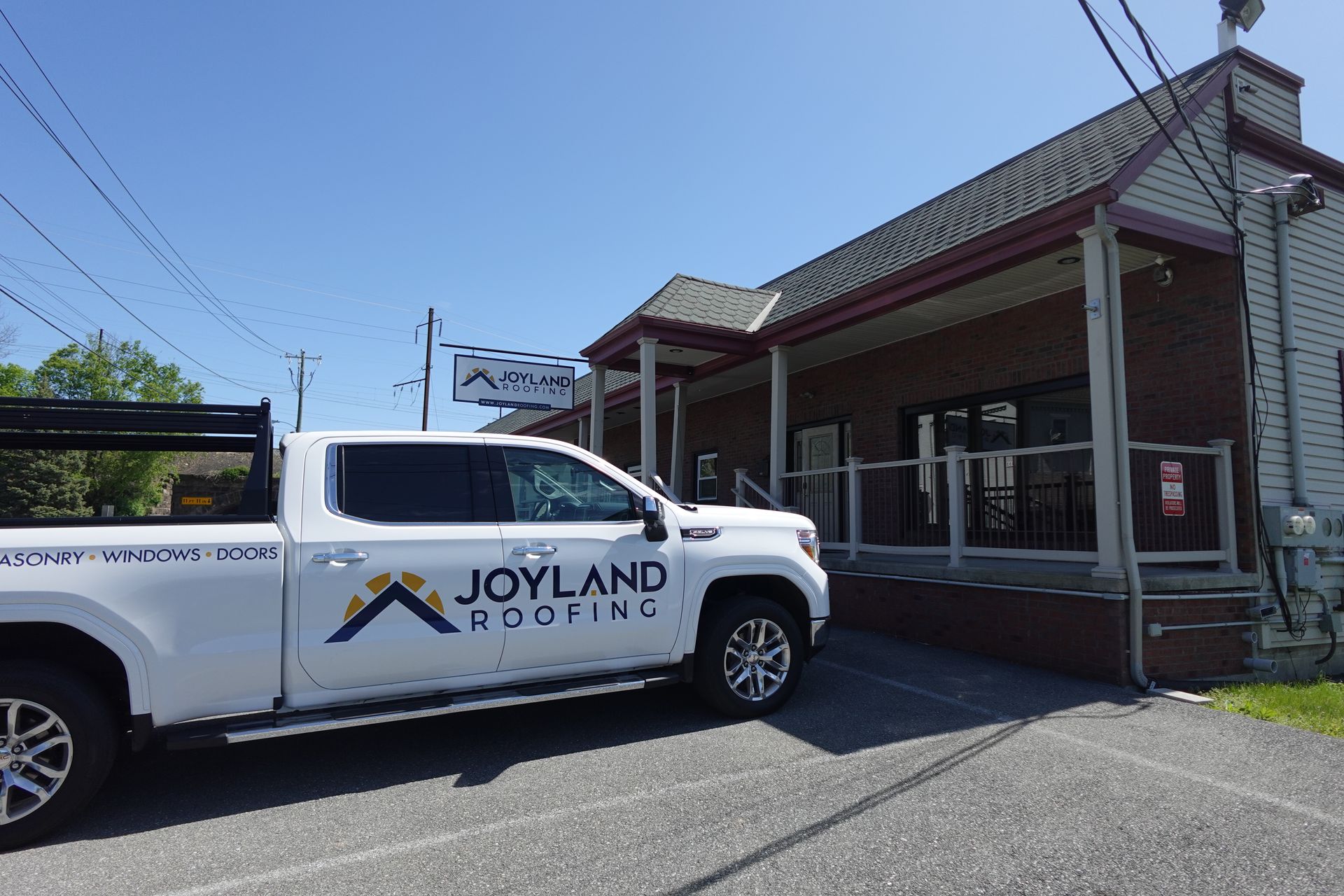 White Joyland Roofing truck parked outside a brick building with a sign on a sunny day.
