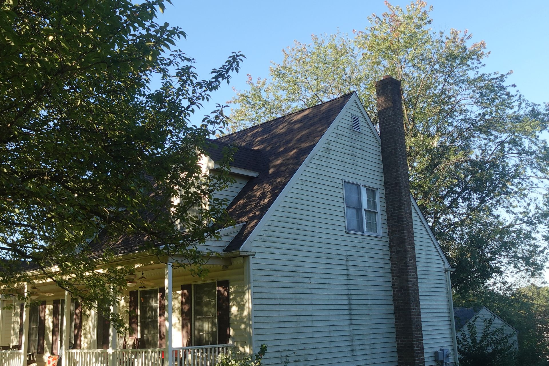 White house with steep roof and chimney, partly shaded by trees in a sunny yard