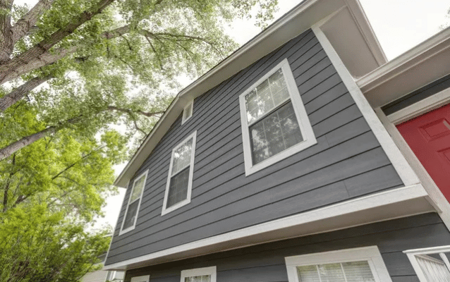 Gray house exterior with white-framed windows, red door, and surrounding tree branches against a cloudy sky.