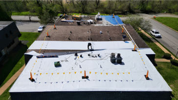 Workers on a flat roof replacing roofing material. Safety cones and debris scattered. Daytime, outdoors.