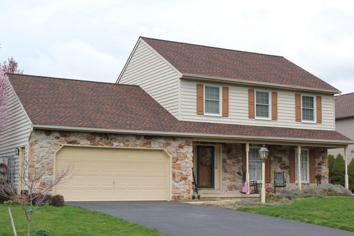 Two-story house with stone facade, beige siding, brown roof, and garage. Driveway and lawn in front.