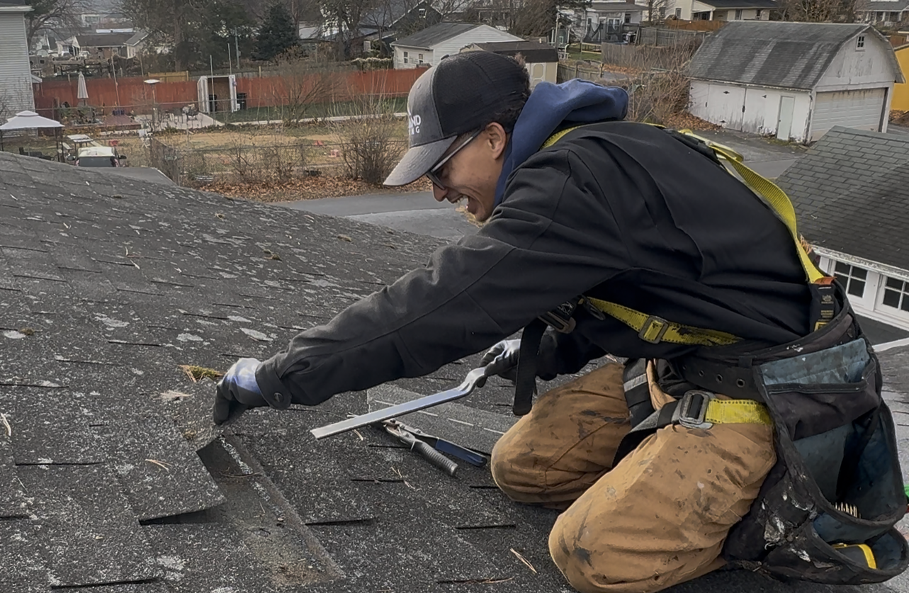Roofer in safety gear kneeling on a rooftop, smiling while working on the shingles.