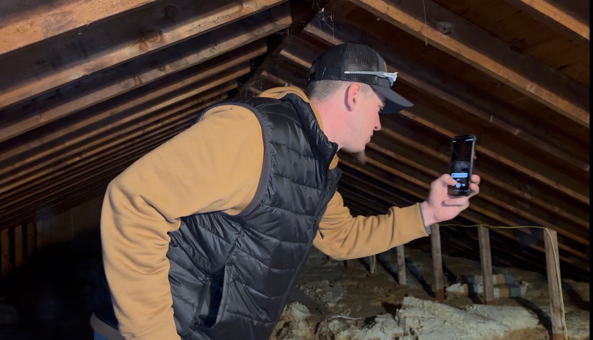 Person in a tan jacket and black vest holding a phone inside a dim attic with exposed rafters.