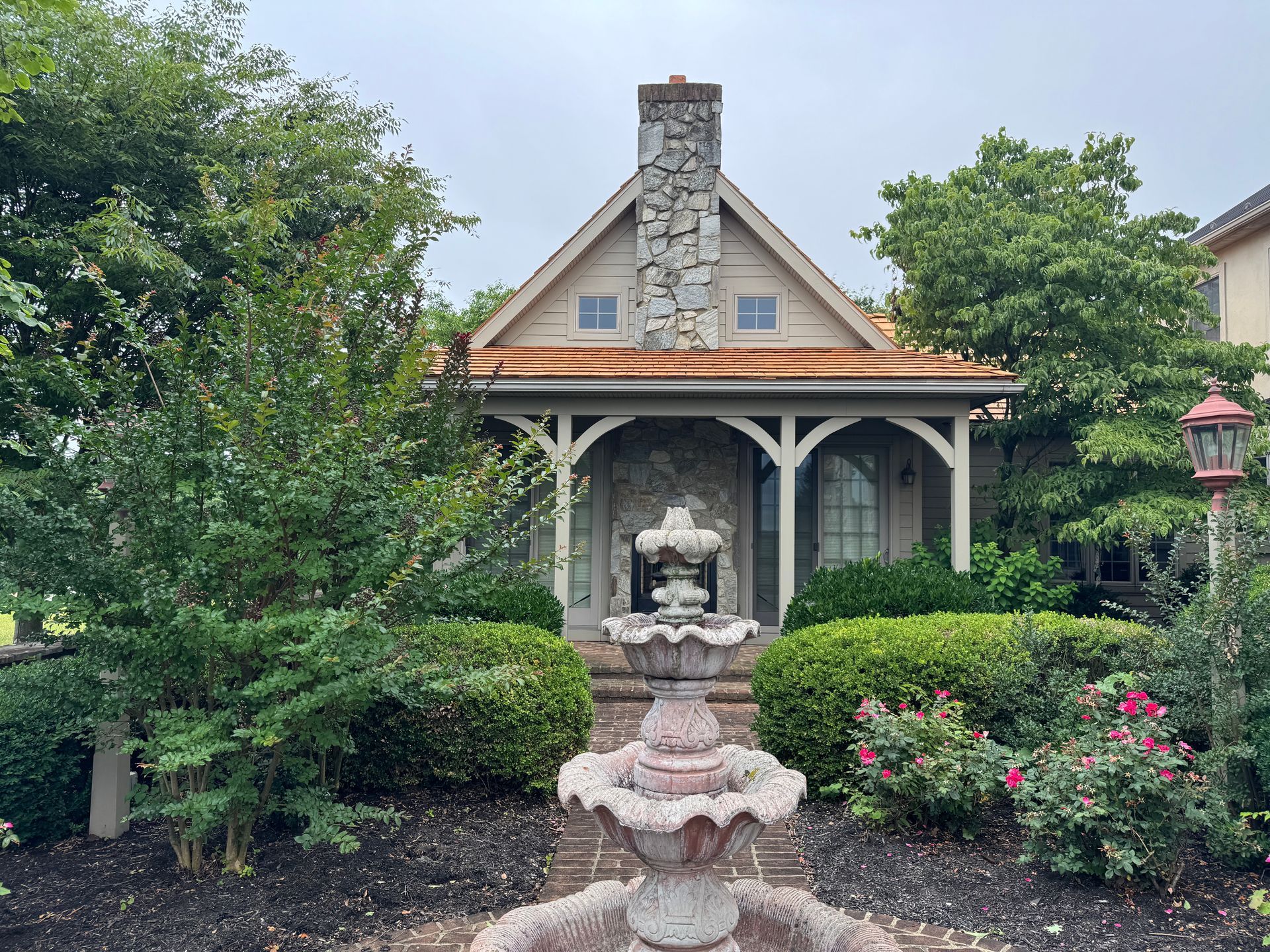 Stone fountain in a lush garden facing a small house with a covered porch and chimney