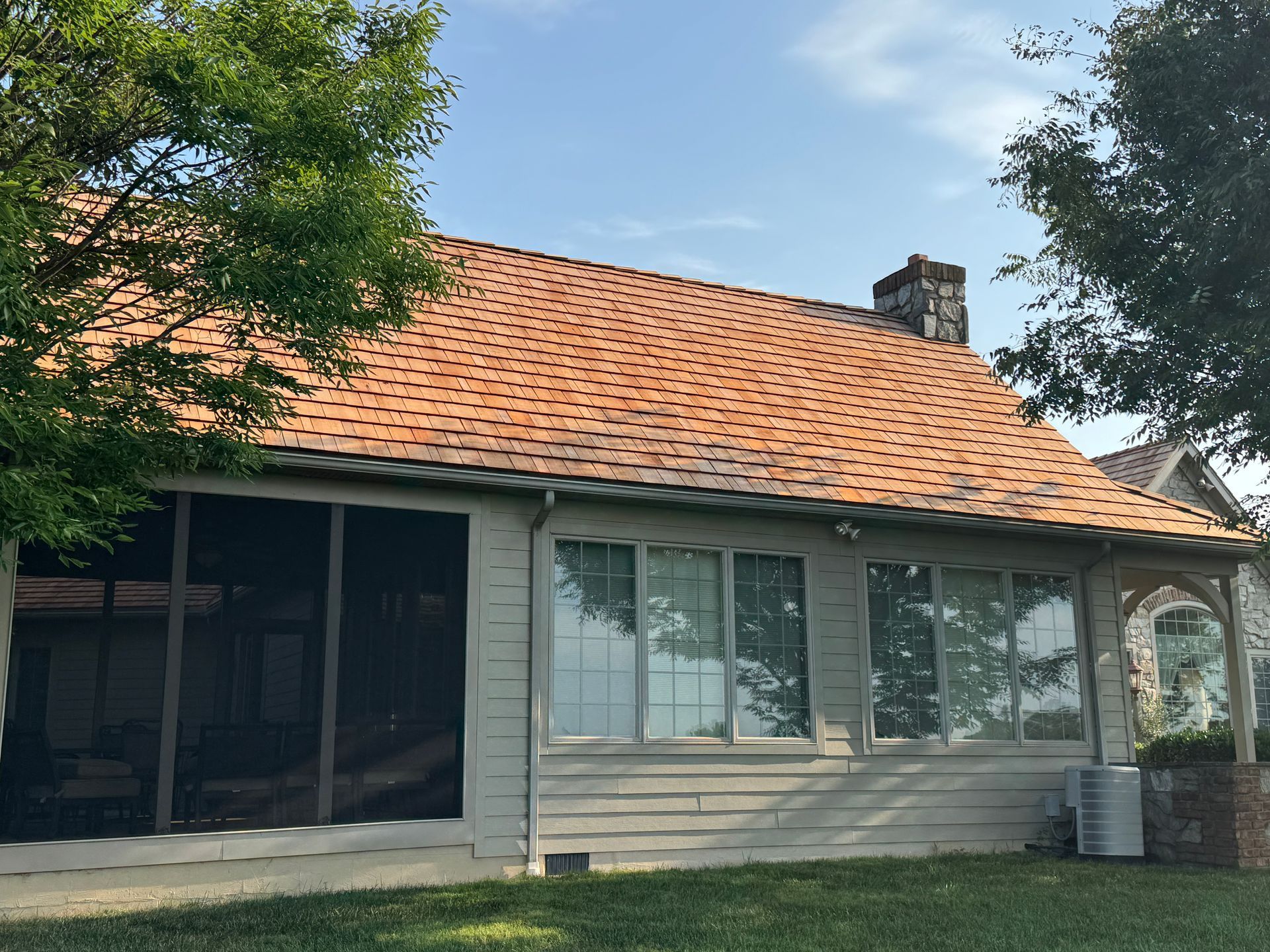 Single-story house with cedar shingle roof, light siding, screened porch, and trees in front