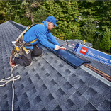 Roofer in blue shirt and hard hat installs roofing material on a slanted roof.