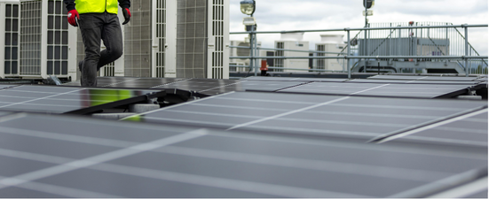 Person in a yellow vest walking on a roof with solar panels. Buildings and overcast sky in background.