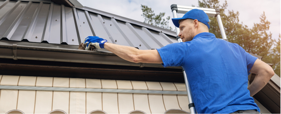 A man on a ladder cleaning a roof gutter. He is wearing blue and using gloves.