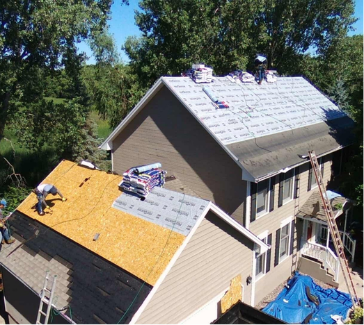 Roofers working on a house with brown siding; shingles and materials visible on the roof.
