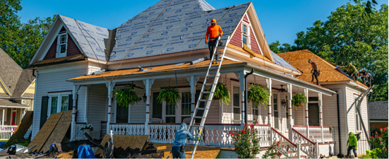 Workers repairing a house roof, one on a ladder, another on the roof. The house is white with a porch.