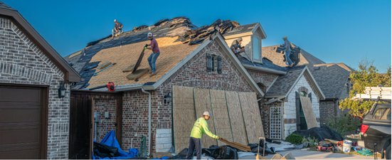 Roofers replacing a damaged roof on a house with brick siding, working on a sunny day.