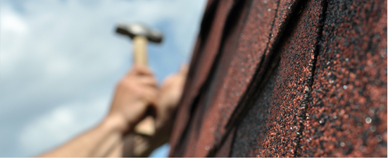 A person hammering shingles on a roof against a cloudy sky.