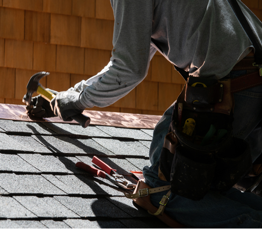 Roofer hammering on a shingled roof, tools in belt, working in daylight.